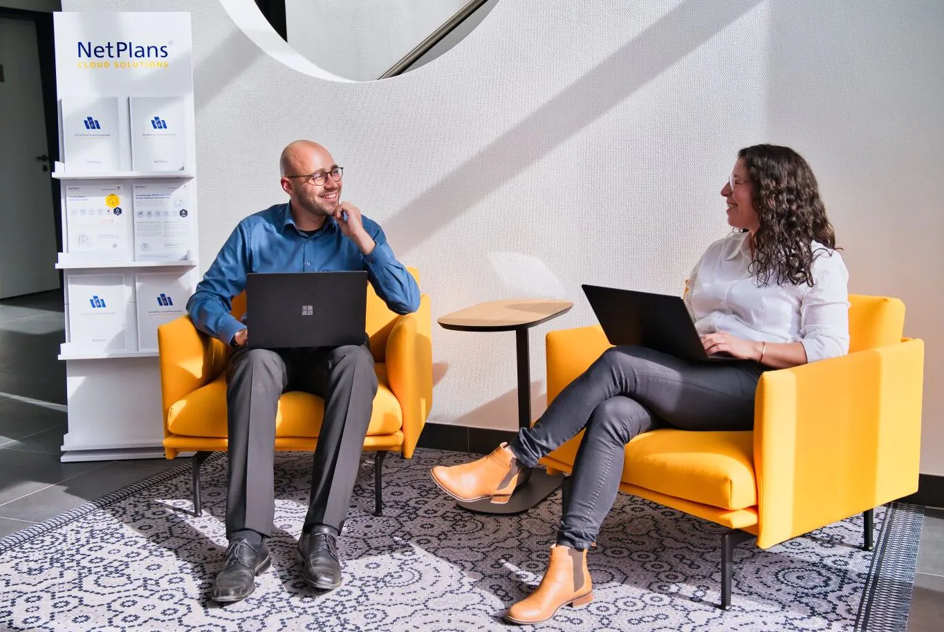 zwei arbeitende und unterhaltende Mitarbeitende mit Laptop sitzen auf gelben Sesseln